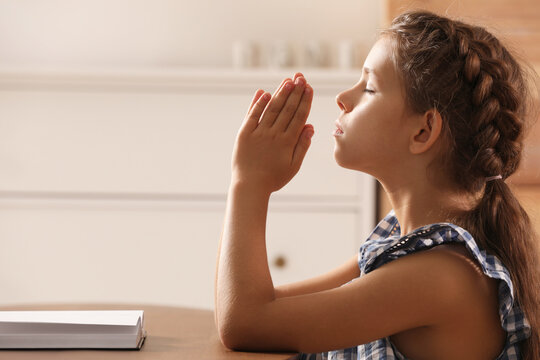 Cute little girl praying over Bible at table in room