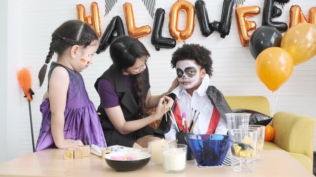 Young woman make-up artist painting vampire face  black skined child for halloween party with little girl in witch dressed.