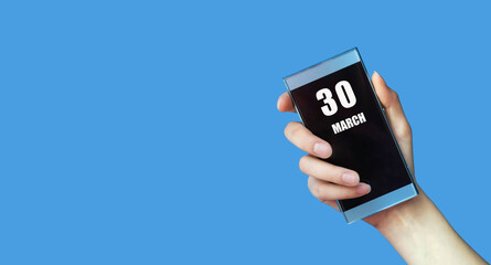 march 30. 30th day of the month, calendar date.Woman's hand holds mobile phone with blank screen on blue isolated background.Spring month, day of the year concept
