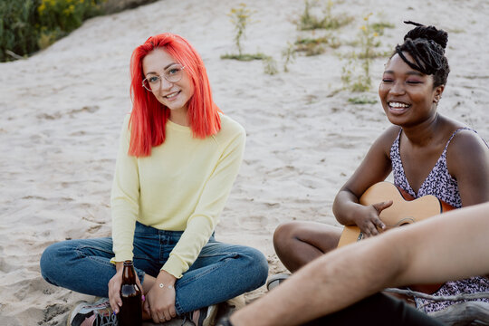 Two women of different nationalities skin colors spending time together on bonfire sitting on sand next to each other african american woman is playing guitar girl with pink hair is drinking beer