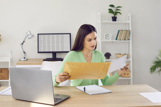 Woman opens envelope that she received by mail. Young girl sitting at desk and holding paper letter she got by post. Beautiful lady reading notification while sitting at table with notebook PC at home