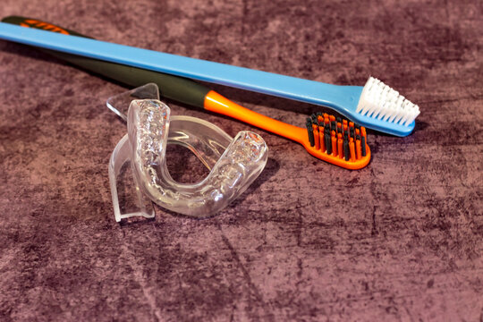 on a lilac background there is adental  mouth guard and two toothbrushes next to it; a silicone impression of teeth and an unused mold for the treatment of bruxism and against night snoring