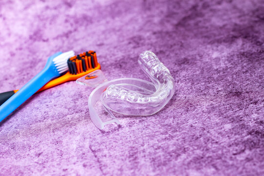 on a lilac background there is adental  mouth guard and two toothbrushes next to it; a silicone impression of teeth and an unused mold for the treatment of bruxism and against night snoring