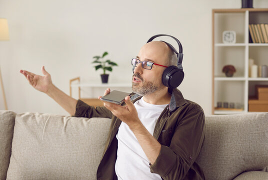 Bald middle aged man in headphones sitting on sofa at home, holding cell phone, talking on speakerphone, recording message, taking audio note, or asking voice assistant to play favorite music station