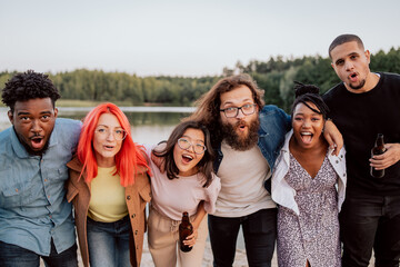 Group of friends huddle close together leaning towards camera drinking beer opening their mouths, laughing, happy, fooling around, scaring each other, a view of the lake in the background