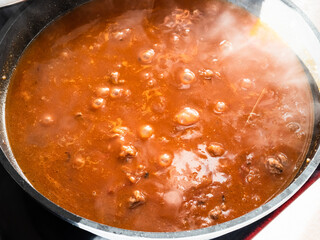 above view of pieces of beef are stewed in red tomato and paprika flavored broth in deep frying pan at home kitchen