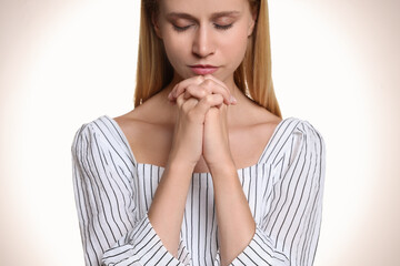 Religious young woman with clasped hands praying against light background, closeup