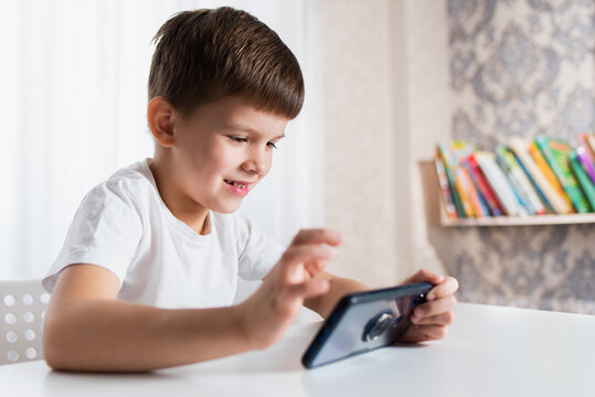A cheerful child in a white T-shirt plays games on the phone at home. Happy boy looks at his smartphone.