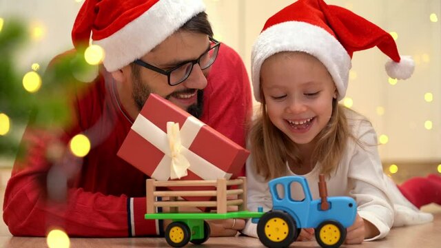 Happy family, father and little girl in red santa hat laugh and play with toy tractor delivering box with new year gift next to christmas tree.