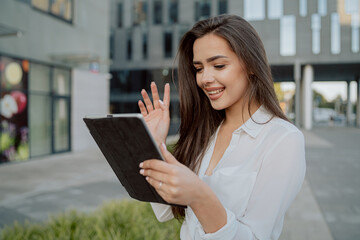 Fototapeta premium Beautiful ambitious businesswoman in white shirt stands in front of glass modern building of insurance corporation company woman holds tablet in hand makes video call, waves to camera, break from work