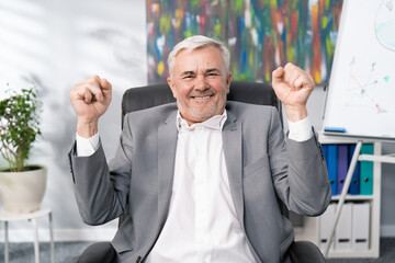 A cheerful smiling businessman in suit with gray hair, elderly, sitting in office chair, raises hands in the air clenched in fists, the head of company won tender, signing contract, victory, happiness