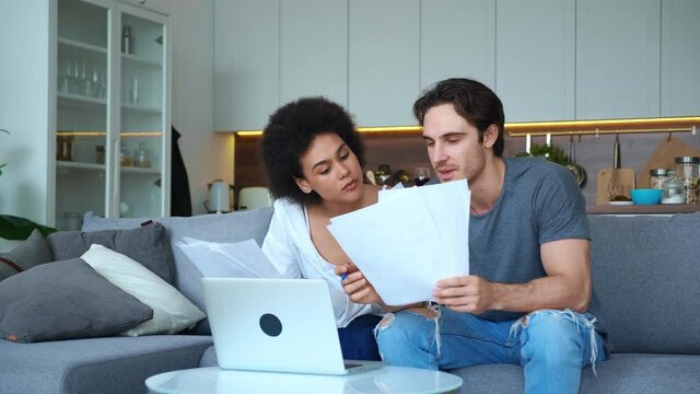 Handsome African American woman and handsome caucasian guy sitting on the sofa in the living room, holding paper sheets of documents and comparing to the contents on the monitor screen, typing text