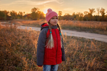 Portrait of pensive preteen girl standing alone among a dry meadow over beautiful autumn sunset. Calm and deep in thoughts. Beauty in nature, outdoor, fall vibes, active family lifestyle