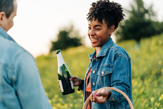 Happy ethnic woman with champagne bottle smiling with crop boyfriend in park