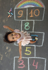 Little African American girl and colorful hopscotch drawn with chalk on asphalt outdoors, top view....