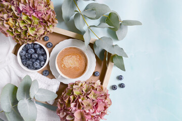 Obraz na płótnie Canvas White cup of cacao, coffee with blueberries, hydrangea flowers, branches of eucalyptus and white blouse on the marble blue background. Vintage morning coffee time. 
