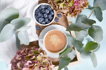 Obraz na płótnie Canvas White cup of cacao, coffee with blueberries, hydrangea flowers, branches of eucalyptus and white blouse on the marble blue background. Vintage morning coffee time. Still life coffee, cacao composition