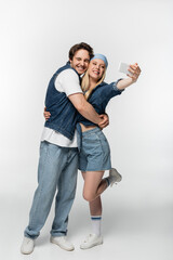 cheerful man with closed eyes embracing woman taking selfie on smartphone on white