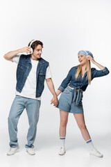 cheerful couple in denim clothing and headphones holding hands while listening music on white