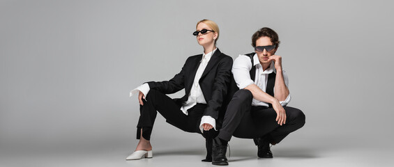 young couple in elegant suits and dark sunglasses posing on grey, banner