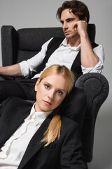 elegant blonde woman looking at camera near blurred man sitting in armchair isolated on grey
