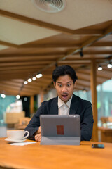 Portrait of shocked Asian businessman in coffee shop using digital tablet computer