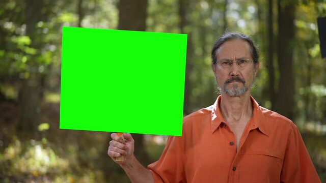 Unhappy, serious mature man holding blank green screen chroma key sign in a forest.