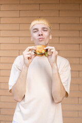 Fast food, leisure, unhealthy eating and people concept - A eating Caucasian man who bitting and holding a burger, wearing a beige T-shirt and looking at the camera on a brick wall background