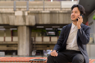 Asian businessman sitting outdoors in Bangkok city using mobile phone