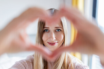 Close up smiling attractive young caucasian woman with blonde hair 30-35 years old showing heart shape gesture at home during coronavirus covid-19 pandemic quarantine and self isolation,
