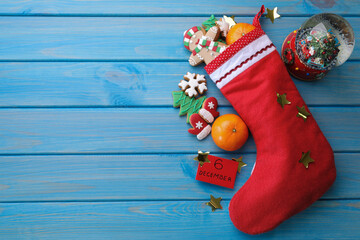 December, 6 - Saint Nicholas Day. Stocking with sweets, paper tag, festive decor and space for text on blue wooden table, flat lay