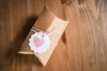Small gift wrapped in craft paper on a natural wooden background. Homemade present with heart shape...