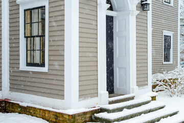 Two vintage double hung windows with a black door on a beige color exterior wooden wall of a house. The windows are dark green with white trim. There's snow on the ground and steps of the entrance.