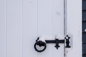 A white wooden door closed with the use of a vintage black wrought iron latch. The mechanism has a round handle. There's fresh white snow on the top of the old handle.  The shed building is white.