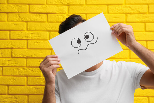 Man hiding emotions using card with drawn frowning face near yellow brick wall