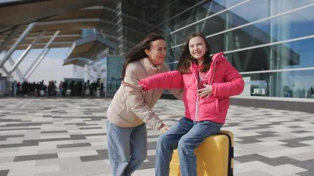 A woman rides her daughter on a suitcase in front of the airport building in anticipation of the trip.