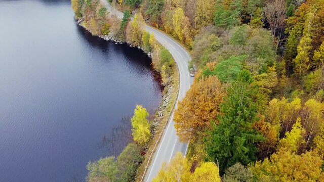 Aerial footage of a curvy road and driving cars.  Drone movie taken from above in Sweden in autumn season. Surroundings with trees and a lake.