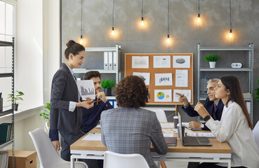 Female employee showing paper graph chat to colleague sitting at briefing desk. Businesspeople discussing project result, startup analysis report together in conference room during meeting at office