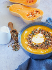 close-up of a pumpkin soup with cream and seeds on top, served in a blue bowl, on the table with an out-of-focus half pumpkin behind it