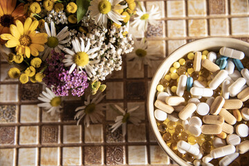 A ceramic bowl with medicinal herbs and a bowl with various pills and tablets are on the countertop. Focus on pills.