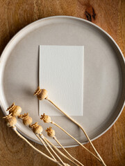 Blank greeting card, invitation and dried flowers on beige ceramic plate and round wooden tray in morning sunlight, flat lay, top view