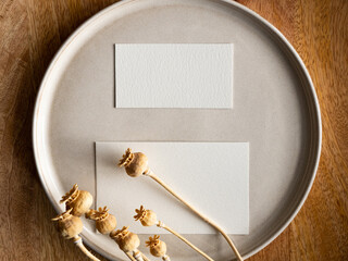 Blank greeting card, invitation and dried flowers on beige ceramic plate and round wooden tray in morning sunlight, flat lay, top view