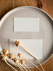 Blank greeting card, invitation and dried flowers on beige ceramic plate and round wooden tray in morning sunlight, flat lay, top view