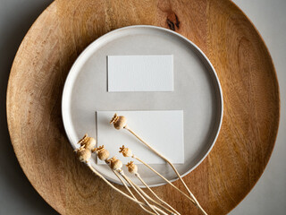 Blank greeting card, invitation and dried flowers on beige ceramic plate and round wooden tray in morning sunlight, flat lay, top view
