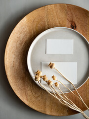 Blank greeting card, invitation and dried flowers on beige ceramic plate and round wooden tray in morning sunlight, flat lay, top view