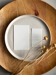 Blank greeting card, invitation and dried flowers on beige ceramic plate and round wooden tray in morning sunlight, flat lay, top view