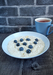 oatmeal porridge with banana, coconut milk and blueberries lies in a gray plate next to a mug of tea on a wooden table against a brick wall. side view. gluten-free and lactose-free vegan breakfast
