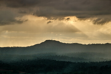 Sunset over the mountains in cloudy sky with sun rays down to the hills with green forest and agricultural field