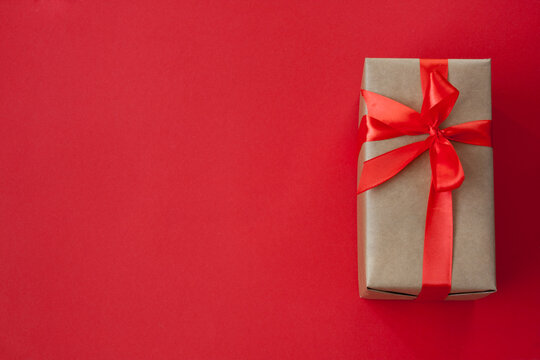 One rectangular gift box, packed in craft paper, tied with red ribbon with bow. Lies on the right on red background. View from above. Flat lay