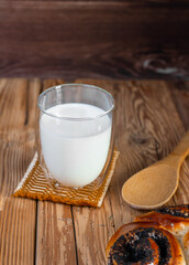 A glass of cold milk with a spoon and a bun with poppy on a wooden table